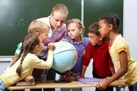 The picture depicts a teacher and five children in a classroom standing and looking at a globe.